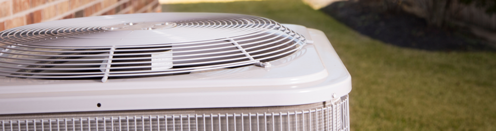 Close-up view of the top portion of an outdoor air conditioning unit, highlighting the fan and protective grille. The surrounding green lawn and brick wall provide a residential backdrop, emphasizing the role of well-maintained and energy-efficient AC systems in promoting environmentally friendly cooling solutions for homes.